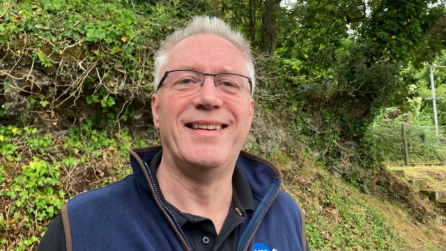 Jeremy Rennick stood by trees. He has grey hair and is wearing a blue fleece.