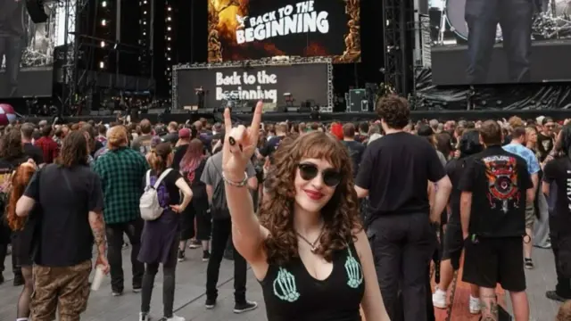 Jessica has long curly brown hair. She’s wearing sunglasses, a black vest with skeleton hands on it and a black shirt. She holds her hand up with the rock and roll hand gesture. She’s in front of a giant stage with “back to the beginning” branding at Villa Park Stadium.