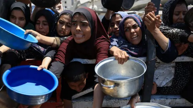 Palestinians gather to receive food from a charity kitchen, amid a hunger crisis, in Gaza City. They are holding out empty bowls and pots. Many of the women are wearing head scarves and look visibly upset as they reach out their arms for food.