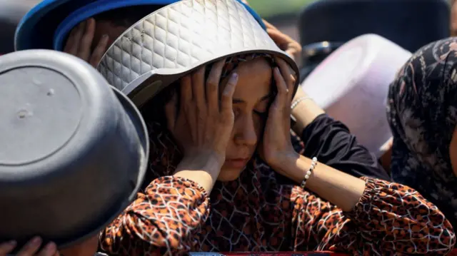 A woman with a metal pot covering her head holds up her hands to the sides of her face with her eyes closed. Around her are other people also holding up plastic basins as they wait to collect aid