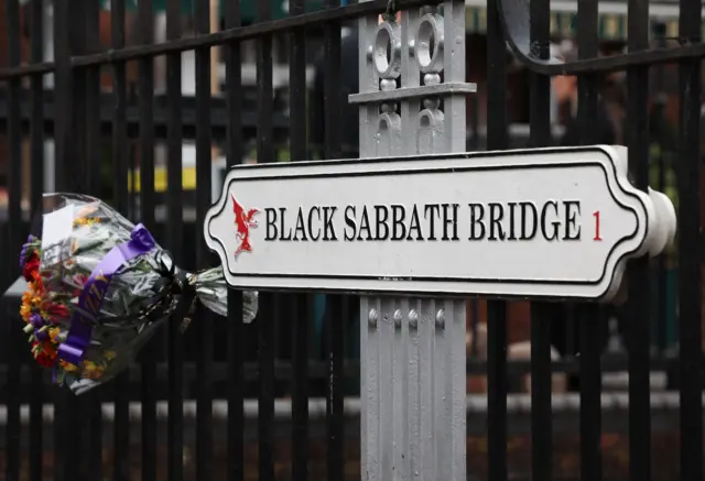 A white sign reads Black Sabbath Bridge. There are black railings behind the sign with a bouquet of flowers attached to them.