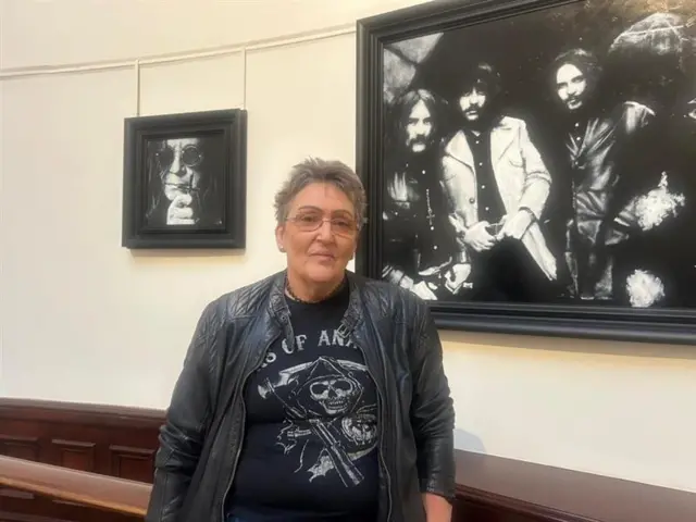A woman with short grey hair and glasses looks at the camera. She is wearing a black t-shirt with the grim reaper on it and a black leather jacket. She stands in Birmingham Museum and Art Gallery in front of a black and white portrait of a young Black Sabbath.