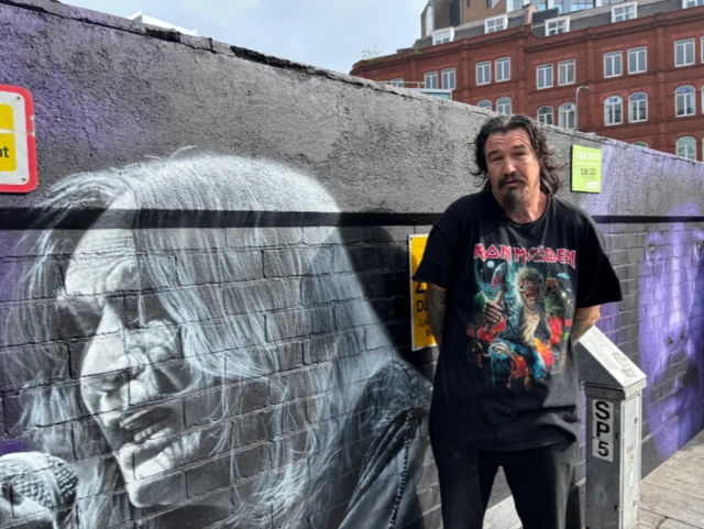 Phillip Clarke stood next to a mural of Ozzy Osbourne. He is wearing a black Iron Maiden t-shirt with black trousers. He has long black hair and a black goatee beard.