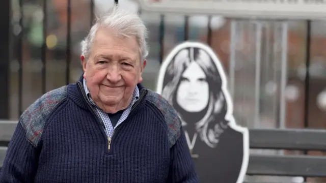 Jim Simpson pictured on the Black Sabbath bench in Broad Street, Birmingham. Behind him is a Ozzy Osbourne cutout in black and white. Mr Simpson is wearing a navy blue fleece with a blue and white checked shirt underneath. He has white hair and is clean shaven.