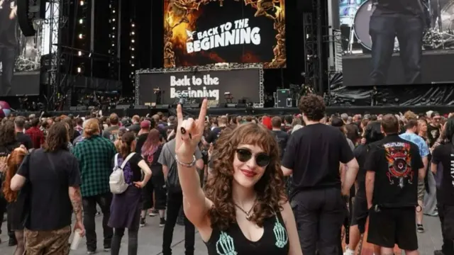 Jessica holding her hand with a rock hand gesture with a black vest on. She has long brown curly hair and is wearing sunglasses. She's at Villa park at the Back to the Beginning concert right at the front.