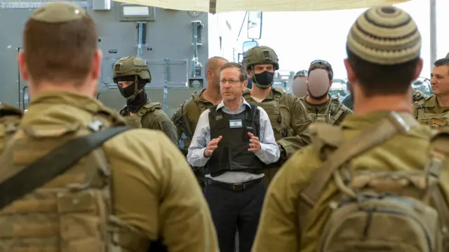 Israeli President Isaac Herzog (central) speaks to members of the IDF during a visit to Gaza. He's standing, his arms partially raised as he speaks wearing a light blue shirt, bulletproof vest with a Israeli flag patch in the front and black slacks