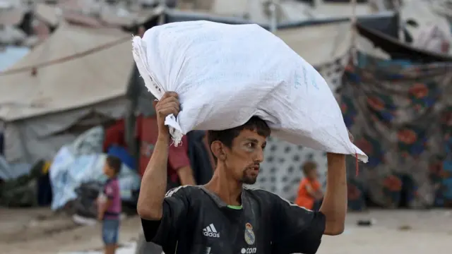 A Gazan man in a dark football jersey with the Adidas logo on the top right side carries a sack of humanitarian aid on his head. A child and a woman can be seen in the blurred background next to a tent encampment