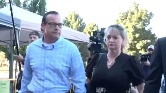 Two people hand in hand looking downcast walking, with lots of reporters following them with cameras