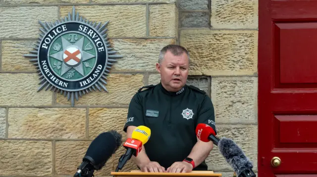 A man in a green PSNI uniform is standing at a brown lectern speaking to people off camera.