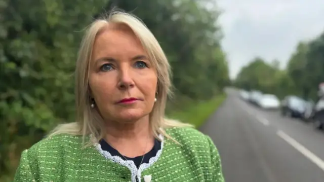 A woman with short blonde hair stares at the camera. She is wearing a black top and green cardigan. She is wearing makeup with dark pink lipstick.