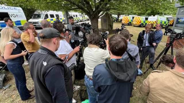 A large group of journalists holding cameras, phones and microphones, gathered on a grassed area. In the background are lots of parked police vans.