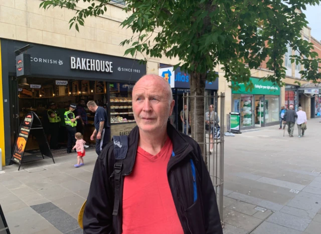 Paul looks away from the camera with a worried expression on his face. He is wearing a red top and a black fleece, and there is a row of shops behind him.