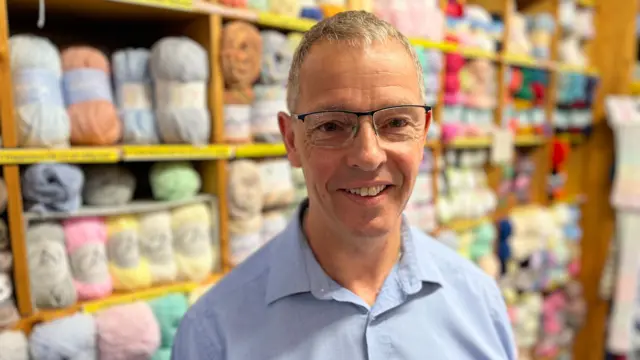 Roger, who wears a light blue shirt and square glasses, smiles at the camera. He is standing in front of large shelved full of balls of yarn.