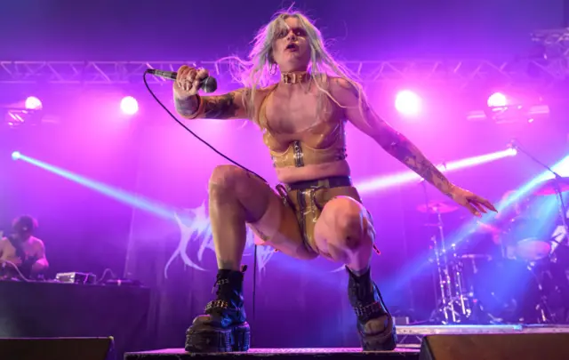 Mimi Barks on stage at Bloodstock Festival with bright pink lights. She's crouched down with big black boots. She's wearing a yellow vest and shorts. She's holding a microphone and staring out into the audience.