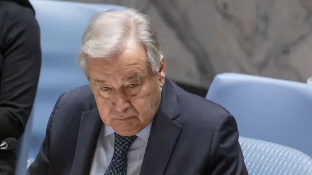 UN Secretary General Antonio Guterrez in dark suit, light blue shirt and tie sits down on blue chair inside UN main chamber
