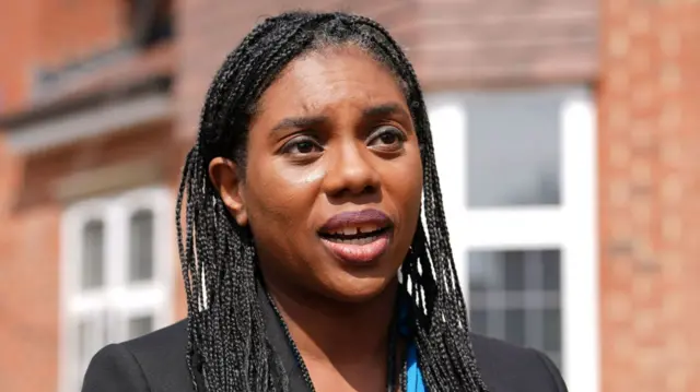 Kemi Badenoch wearing a black suit jacket over a blue top as she talks to the media in front of a house.