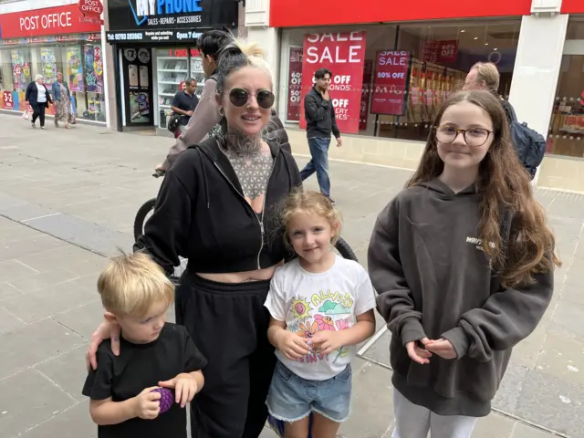 Kylie, who wears dark sunglasses, a black fleece and black tracksuit bottoms, stands with her three children - two girls and a boy.