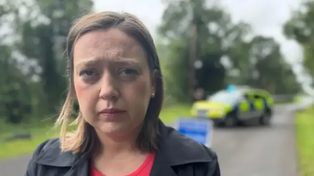 A woman with short brown hair stares at the camera as she stands in front of a police cordon and car. She is wearing a pink top and black coat.