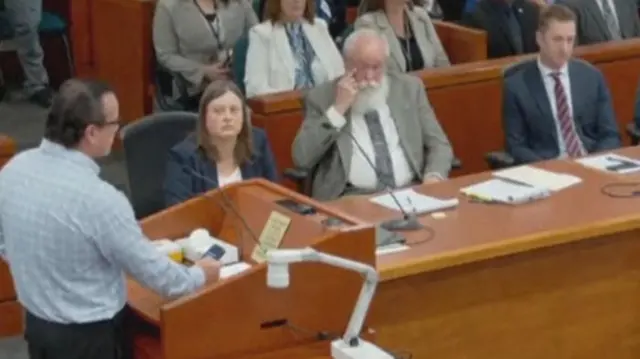 A man at a podium speaking as people in the courtroom sit on benches watching him