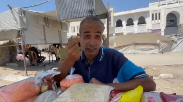 A man gesturing with one hand, while he leans on the other arm on a table, which has pasta, lentils and bagged food on. Behind him, the street is dusty and sandy, with makeshift shelters and a white building in the background