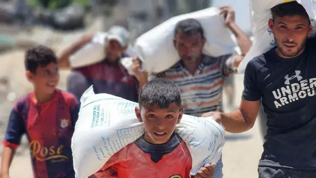 A boy in the centre of the image looks strained as he carries a large heavy white sack on his shoulders. In the background, three men and a boy also carry the sacks, looking laboured