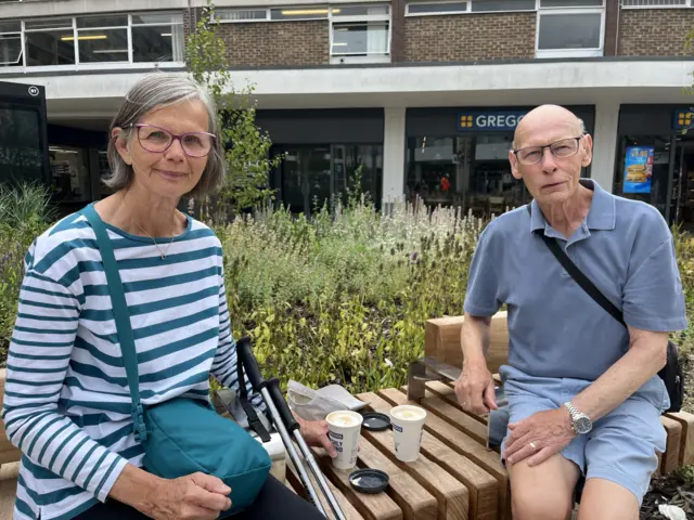Elizabeth and Robert sit on a bench in the town centre with two coffees. There are lots of plants and wildflowers in a planter behind them.