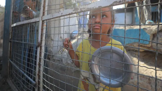 A child holds an empty bowl at a fence in Gaza