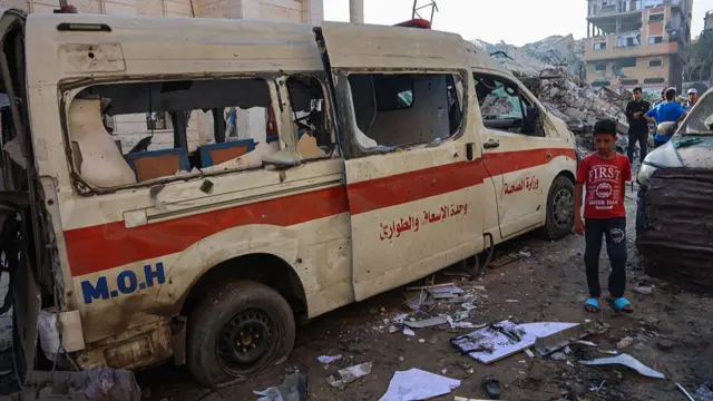 A ruined ambulance sits in a ruined street surrounded by rubble as a boy stands next to it