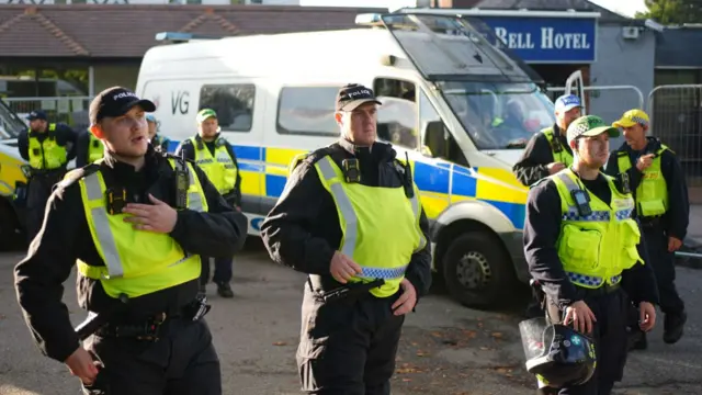 A large group of uniformed police officers standing in front of a large van, which is parked at the entrance to the Bell Hotel.