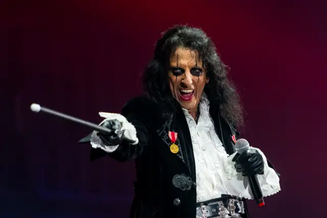 Alice Cooper. He is on stage  wearing his show make up wearing a black  jacket and white shirt. He is holding what looks like a magic wand in one hand and a microphone in the other.