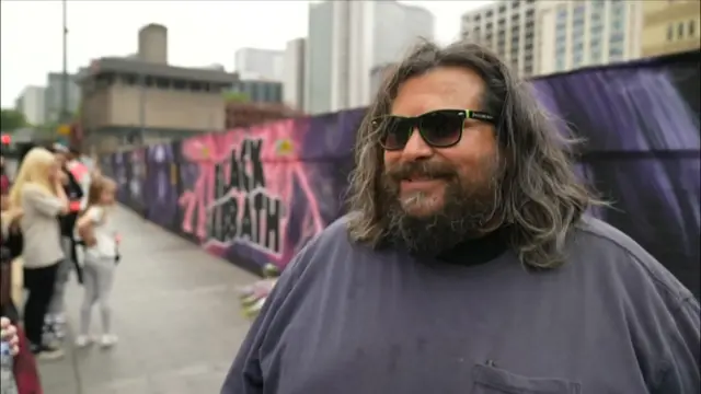 A man with shoulder-length dark hair smiles at the camera. He wears a grey shirt and black sunglasses. The man is stood in front of the Black Sabbath mural in Birmingham city centre, which says Black Sabbath in the middle in bold black lettering with pink electricity bolts around it. The Black Sabbath members are included on the mural - many of them drawn in purple.