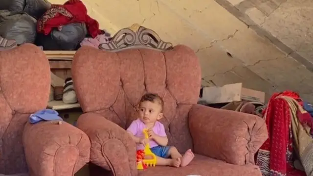 A toddled in a pink shirt and jeans shorts plays on a dust-covered armchair under a ruined ceiling, piles of clothes and debris around him