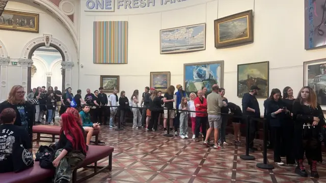 A queue of people stretches right the way across a room in Birmingham Museum and Art Gallery. There are several paintings on the white wall behind them and a black queue barrier is in front of them.