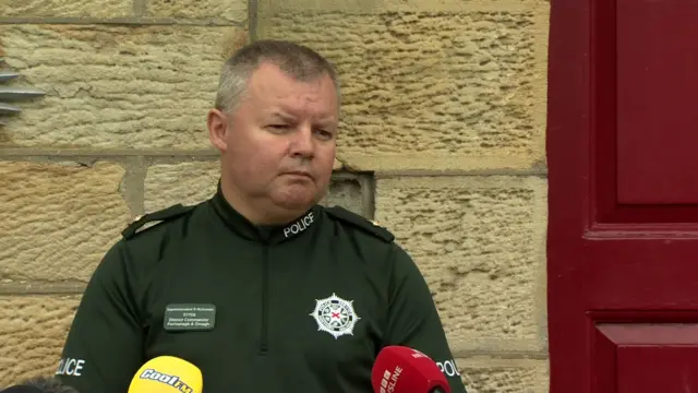 A police officer with short dark and grey hair wears a bottle green uniform and stands in front of mics at a press conference.