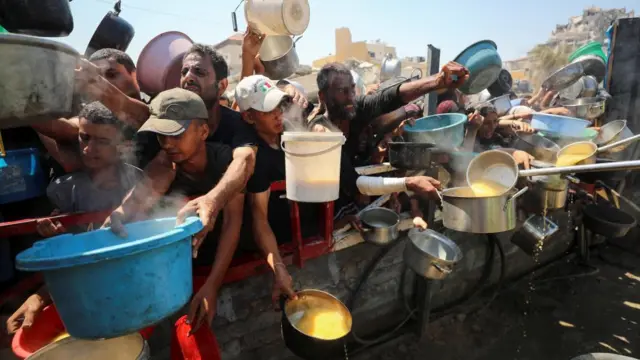 A crowd of people pushed up against a wooden fence leaning over it. They are all holding various pots, pans and buckets, trying to catch liquid that is being poured into them. They look agitated and struggling