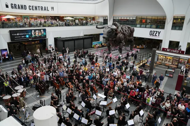 A photo taken from a balcony of an orchestra sitting in a large open-plan concourse with a metal mechanical bull in the background. A crowd of people has gathered.