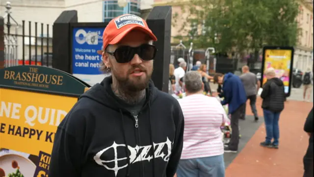 A man wearing black sunglasses, an orange baseball cap and a black hoodie with Ozzy on it in black lettering. He stands in the middle of a Birmingham street with several people behind him