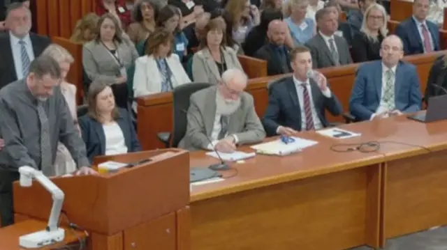 Laramie standing speaking as a busy courtroom watches on