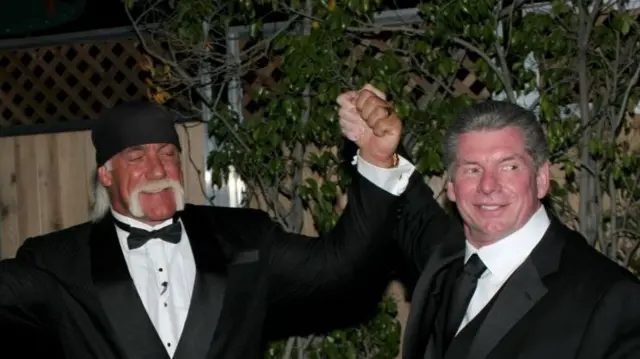 Hogan and McMahon, in suits, hold hands and raise them in the air between them as they smile. They're stood outside at night time. There's a fence and shrubbery behind them