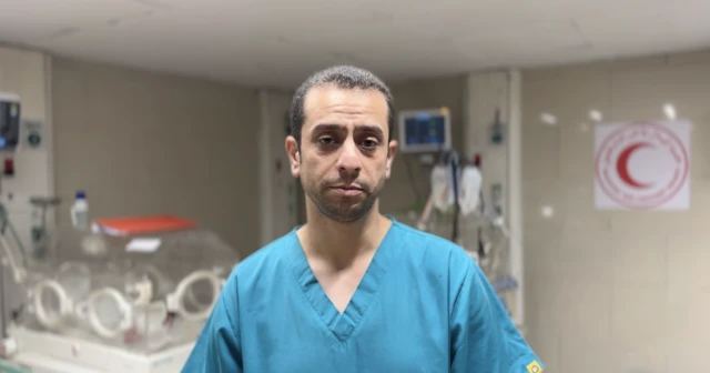 The doctor, wearing blue scrubs and standing in an operating theatre, looks straight down the barrel of the camera, with a serious look on his face.