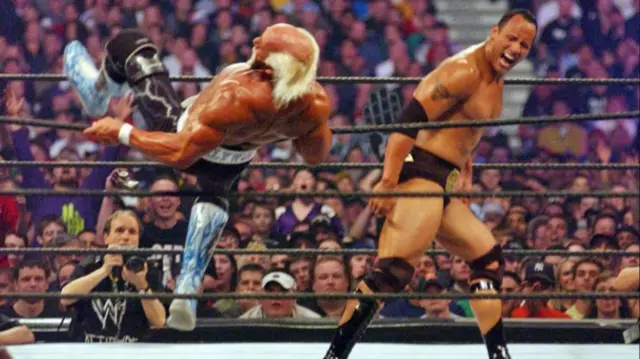 The Rock and Hogan in the wrestling ring. Hogan appears to fall over the ropes as the Rock walks away.