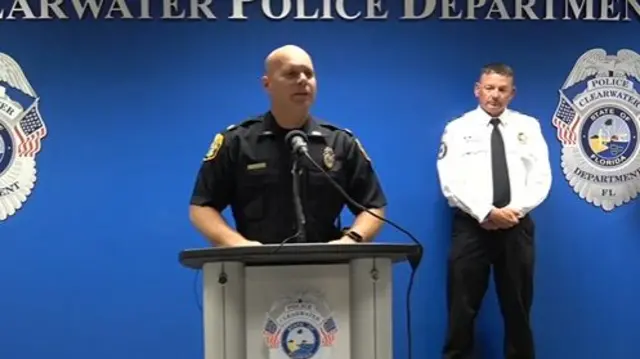 A police officer standing at a podium speaking into a microphone. Another officer stands behind him