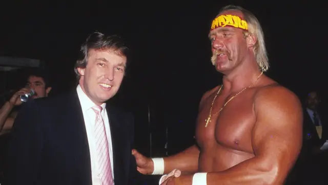 Donald Trump (left) wearing a black suit over a white shirt and pink tie poses shakes hands with a Hulk Hogan who is shirtless and wearing a bright yellow underwear.