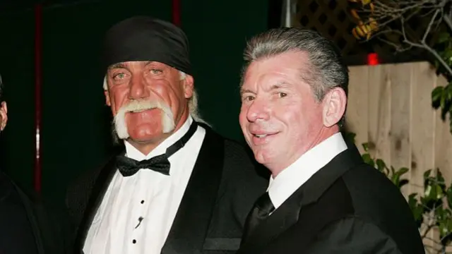 Hogan and McMahon smiling at the camera wearing suits stood outside in the dark in front of a fence