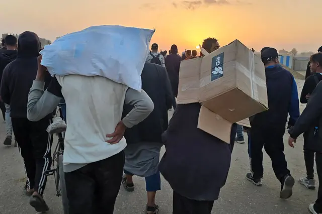 Two men carry aid from a site in Gaza. One box is marked with the GHF logo.