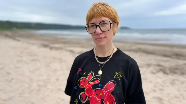 Emma Bailey has short cropped ginger hair, wears two gold necklaces and large clear square glasses with black frames. The sand and sea of Croy Shore, near Turnberry, Scotland, can be see behind her.