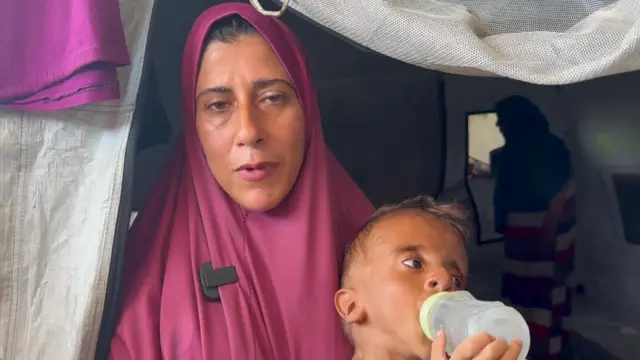 A young woman with her head and body covered by a magenta chador while holding a young child on her lap. The boy is sucking on an empty plastic baby bottle with a green cap