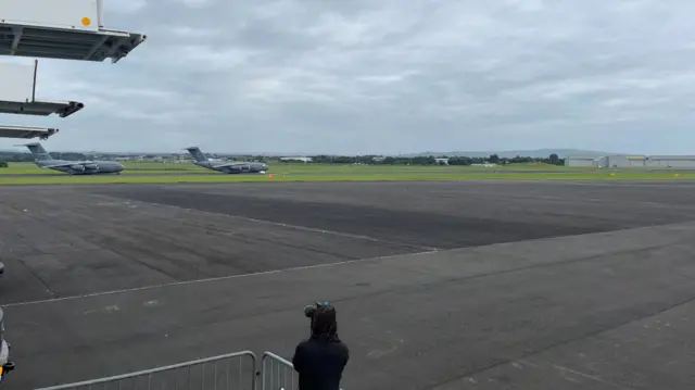 An empty runway at Glasgow Prestwick airport with military planes on the tarmac and green grass and hills in the distance