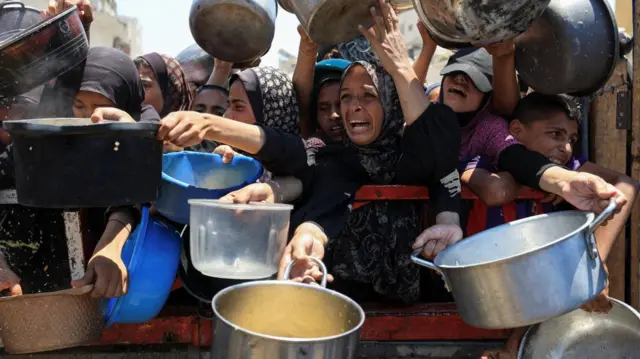 A chaotic scene of Palestinians holding pots and pans, scrambling over each other, as they try to receive food from a charity kitchen.