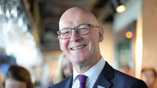 Swinney headshot smiling at the camera. The background is blurry but there are a number of people behind him. He wears glasses and is bald